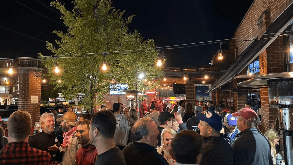 A packed nighttime outdoor patio scene under string lights, with fans in Alabama gear gathered under trees