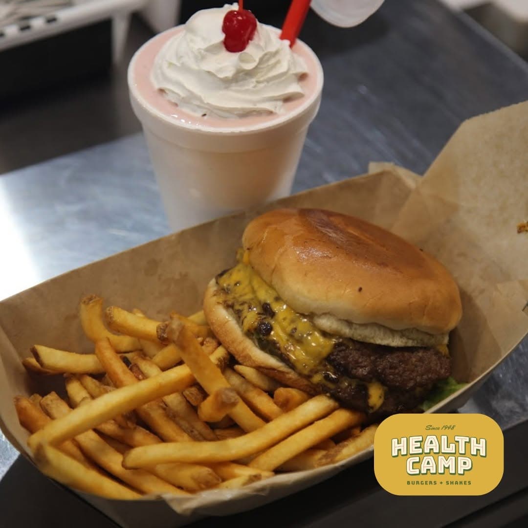 A cheeseburger and fries in a paper tray next to a whipped-cream-topped shake, with the Health Camp Burgers + Shakes logo