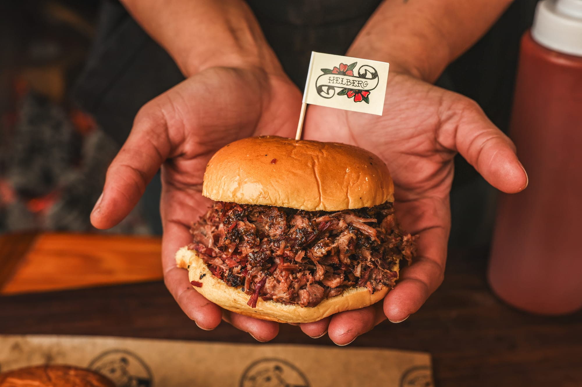 Two hands holding a pulled-pork sandwich topped with a small Helberg flag pick, a bottle of sauce in the background