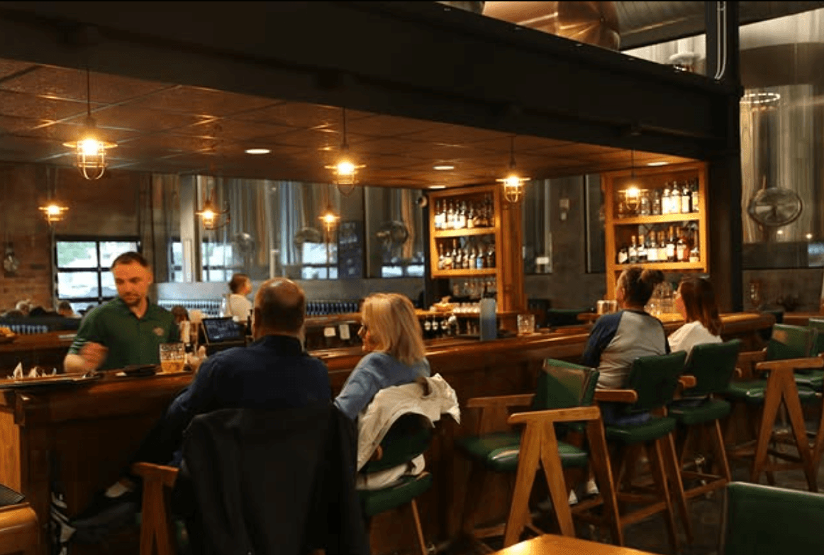 Guests seated at the bar inside Pivovar Taproom, with warm pendant lighting and steel brewing tanks visible behind the bar