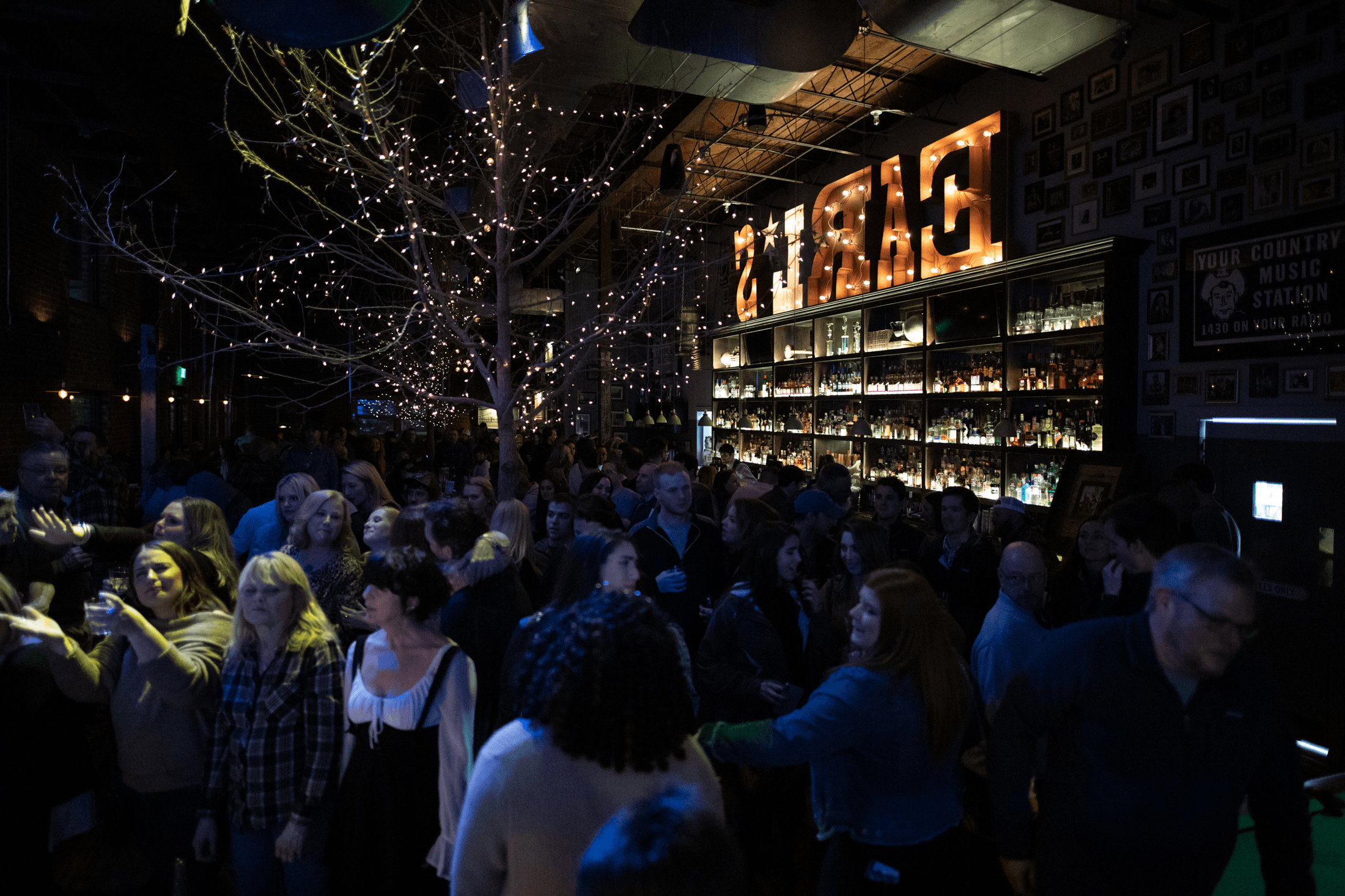 A packed crowd dancing under a string-lit tree at Earl's, with the glowing EARL'S sign and a fully stocked back bar behind