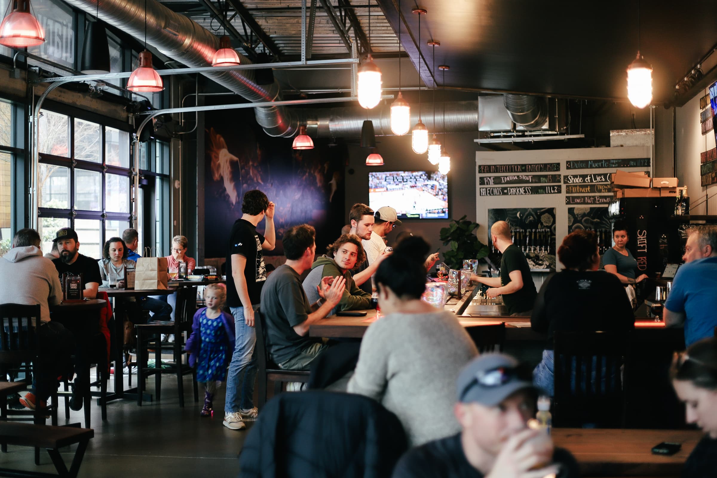 A crowd of patrons at long tables inside Incendiary Brewing Company's industrial taproom, with chalkboard beer menus and a bartender pouring at the tap wall
